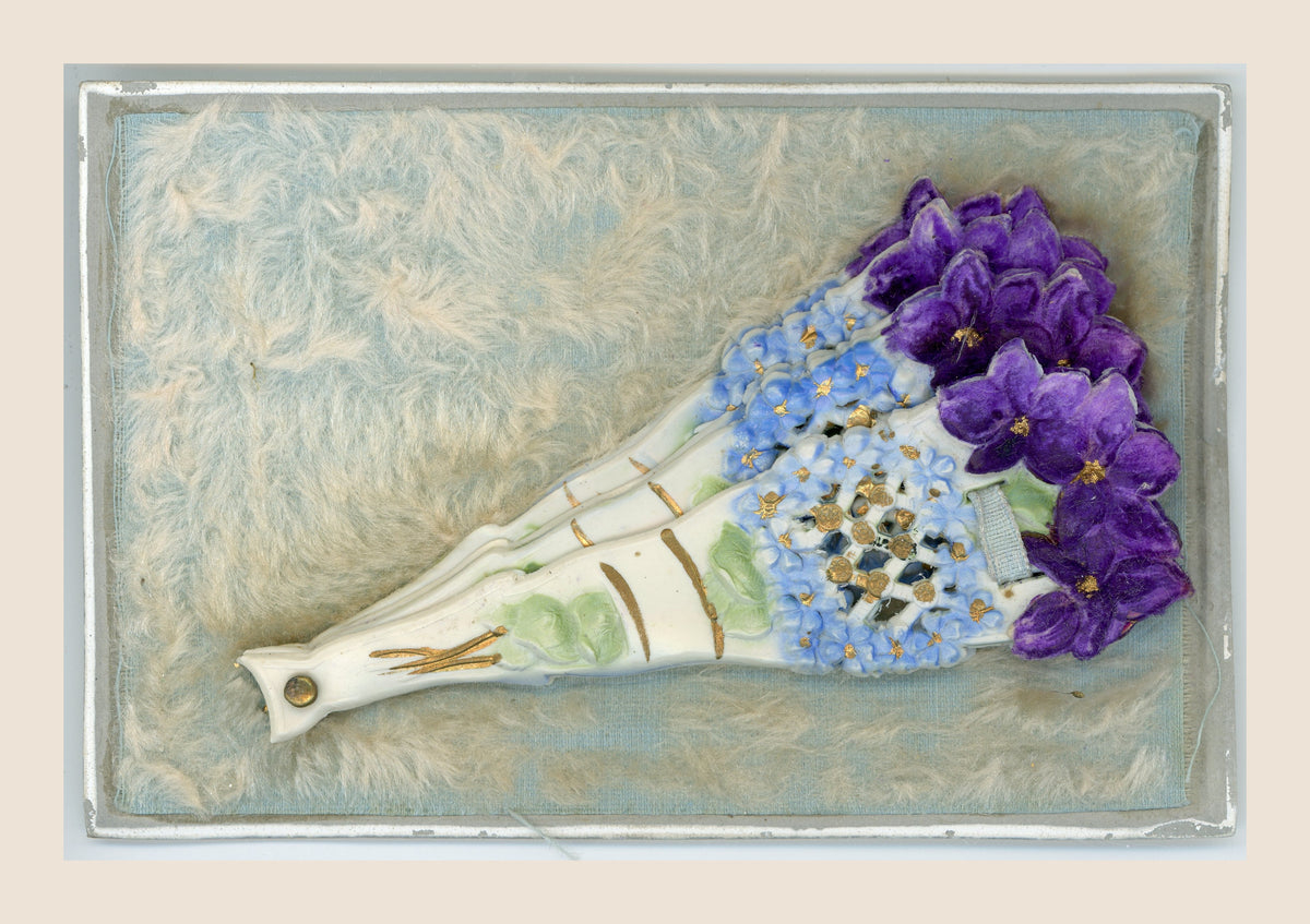 Antique postcard with a closed floral fan on a pale blue textile background covered with soft cream fibres, within a silver-coloured border.