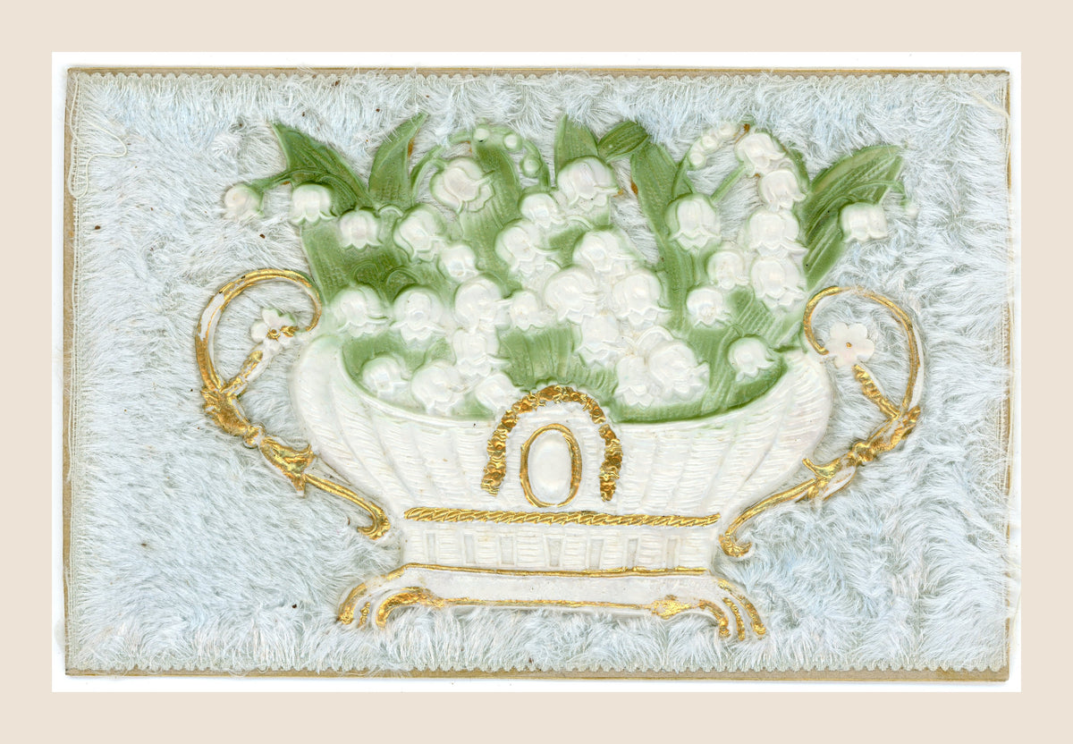 Antique postcard with white lily of the valley flowers in an ornate white and gold jardinière, in relief on a pale blue fibrous textile background.