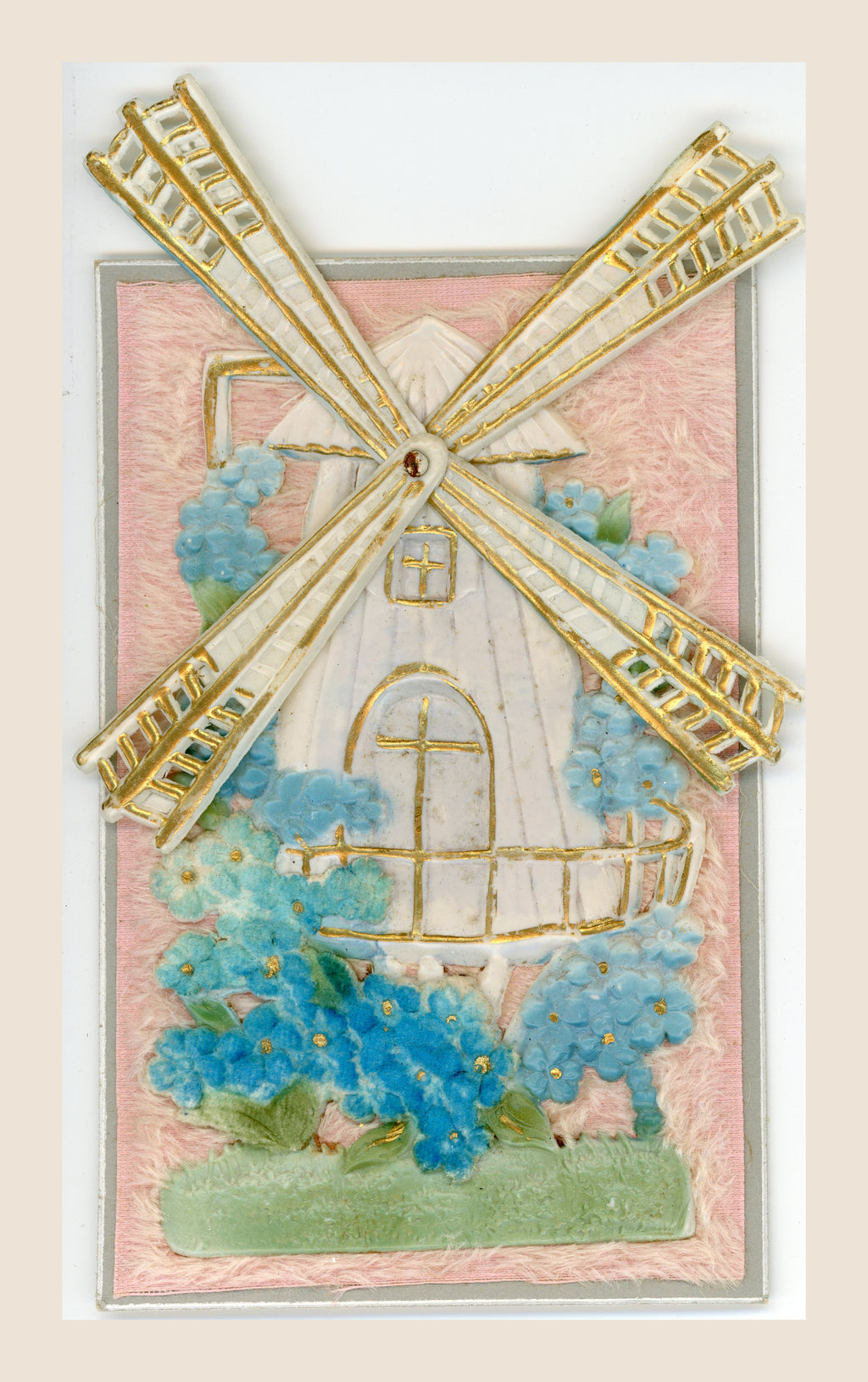 Antique postcard featuring a cream and gold windmill with four movable sails spread out, surrounded by blue flowers on a soft pink textile background.