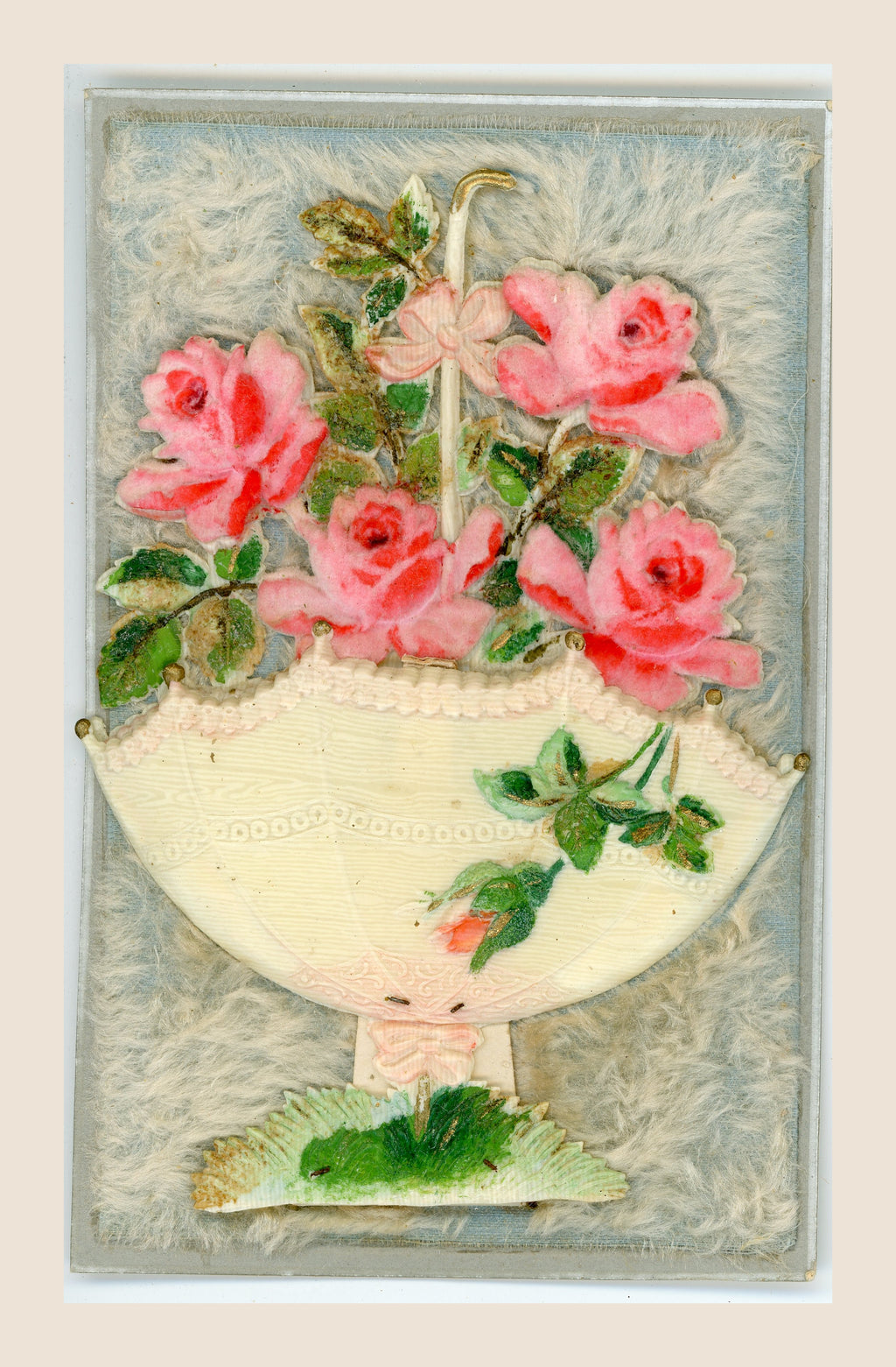 Antique pop-up postcard laid flat, with four pink fabric roses, an upturned cream parasol and green foliage on a pale blue, fibrous textile background.