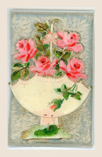 Antique pop-up postcard laid flat, with four pink fabric roses, an upturned cream parasol and green foliage on a pale blue, fibrous textile background.