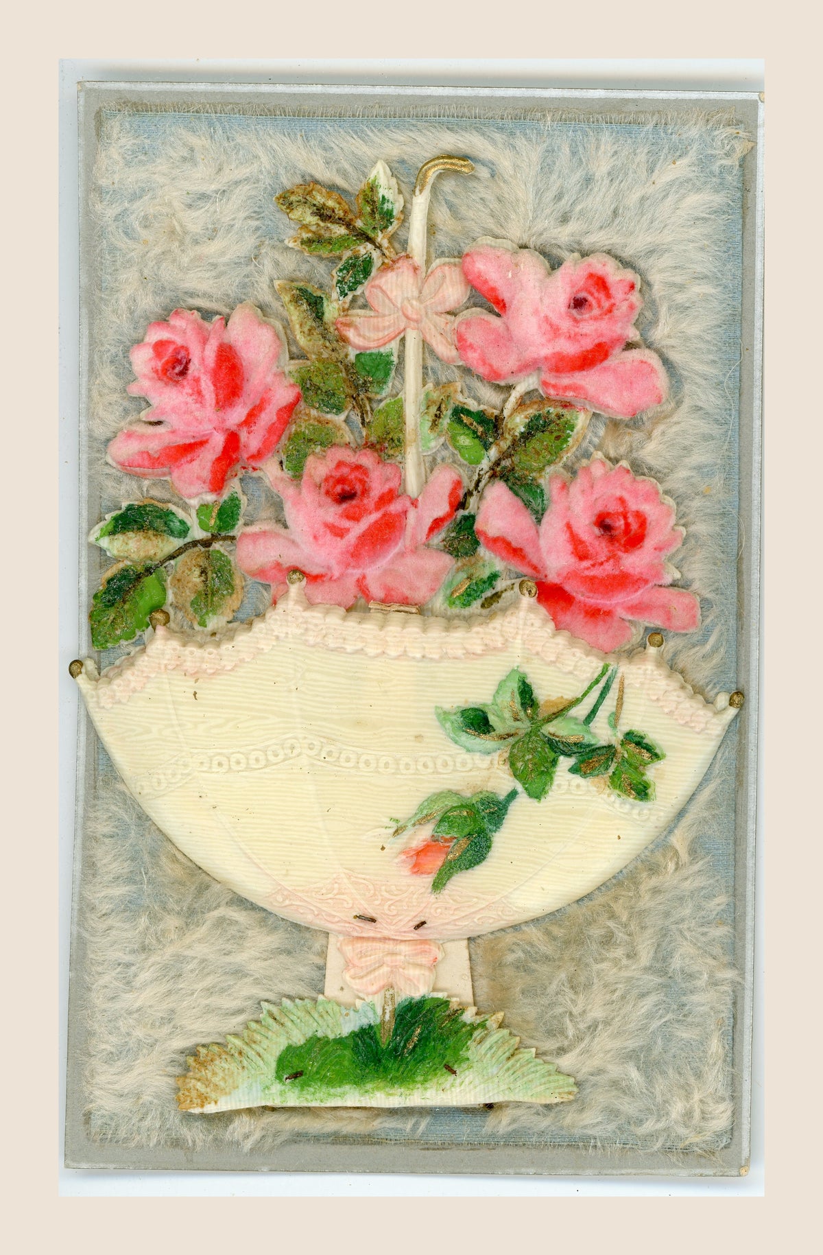 Antique pop-up postcard laid flat, with four pink fabric roses, an upturned cream parasol and green foliage on a pale blue, fibrous textile background.