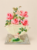 Antique pop-up postcard standing open on a beige background, with pink fabric roses above a cream parasol, green leaves and a rectangular textile-covered base.