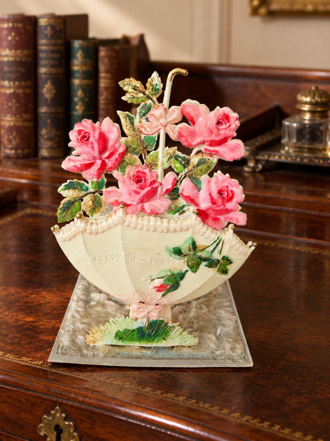 Antique pop-up postcard with pink fabric roses and a cream parasol, standing on a wooden writing desk with old books and an inkwell in the background.