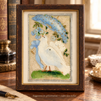 Antique postcard featuring a white dove beneath a blue fabric flower parasol on cream textile, displayed in a patinated wooden frame on a vintage writing desk. Frame not included.