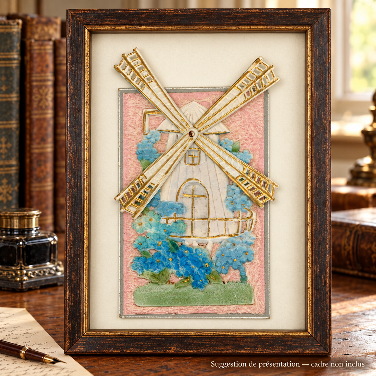 Antique postcard featuring a cream and gold windmill with movable sails spread out, blue flowers and a pink textile background, displayed in a patinated wooden frame on a vintage desk. Frame not included.