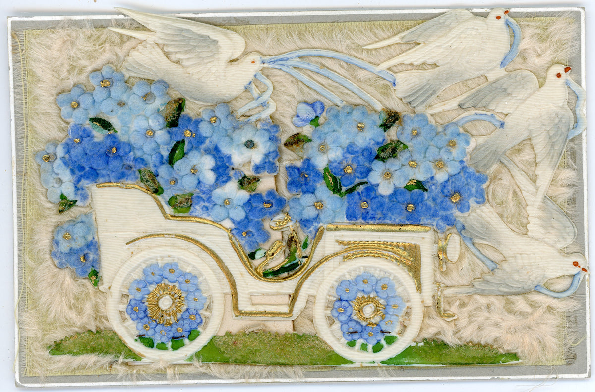 Antique postcard with a cream motor car, blue fabric flowers and four white doves carrying blue ribbons, in relief on a pale fibrous textile background.